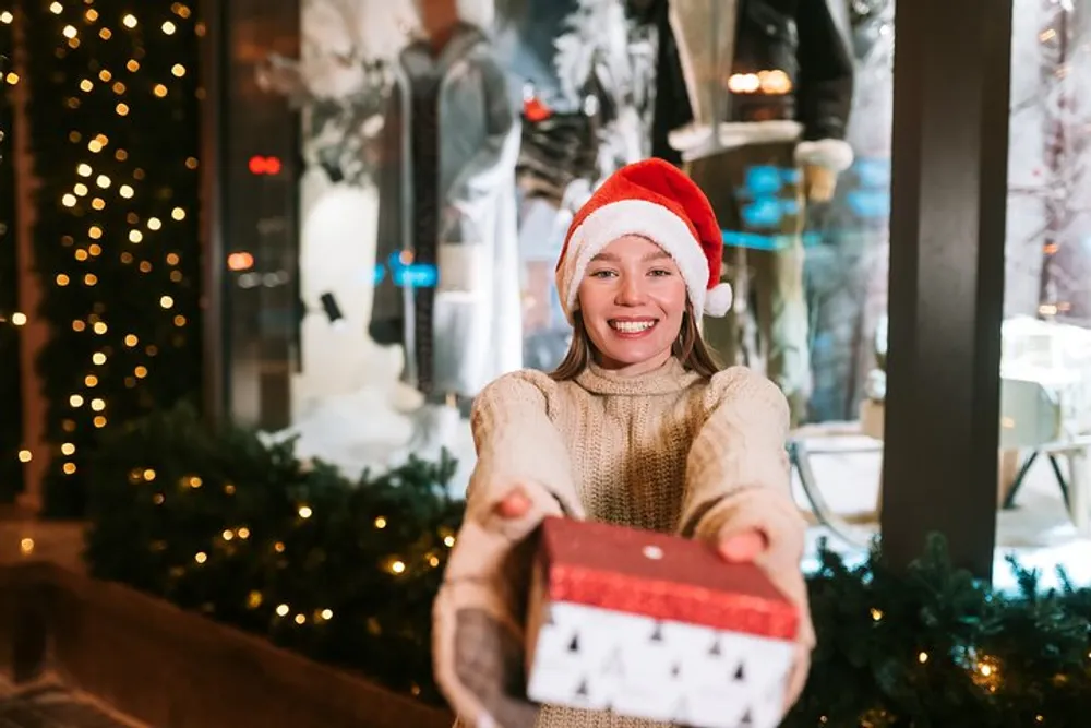 A smiling girl wearing a Santa hat is holding out a Christmas present with festive decorations in the background