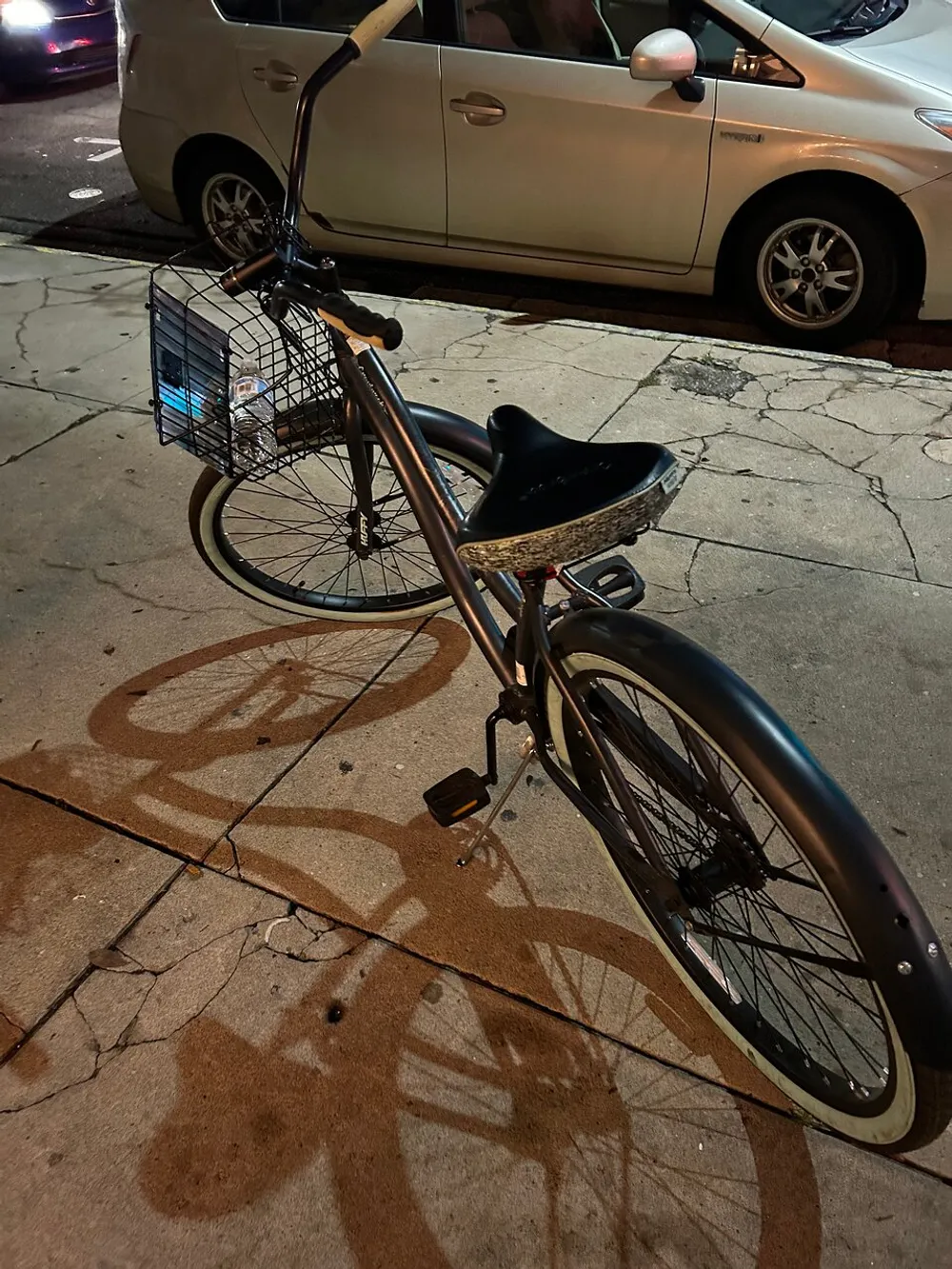 The image shows a single bicycle equipped with a front basket parked on a sidewalk at night casting a shadow under street lighting with a car parked in the background