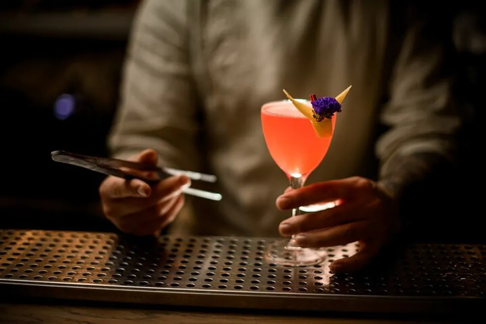 A person is holding a mobile phone in one hand while gently grasping a stem glass with a pink cocktail garnished with an orange slice and a purple flower in the other possibly standing at a bar with a dimly lit ambience