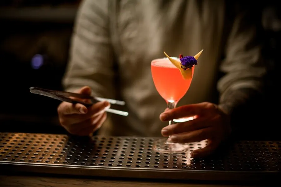 A person is holding a mobile phone in one hand while gently grasping a stem glass with a pink cocktail garnished with an orange slice and a purple flower in the other, possibly standing at a bar with a dimly lit ambience.