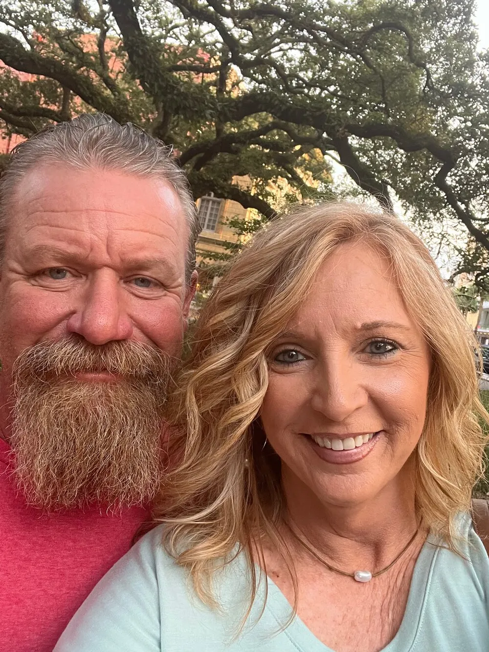 A man with a beard and a woman with blonde hair are smiling for a selfie with a large tree and buildings in the background