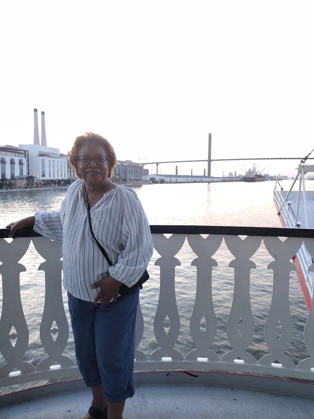A smiling person stands by a railing on a waterfront with a bridge spanning the water in the background during what appears to be either sunrise or sunset