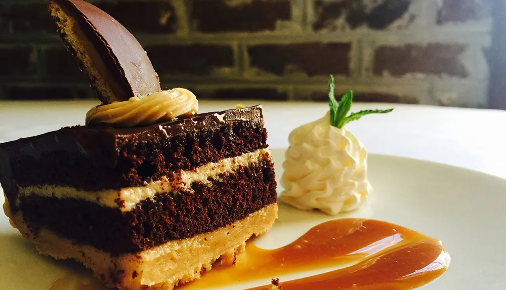 A slice of layered chocolate cake with frosting is presented on a plate with caramel sauce accompanied by a swirl of cream garnished with a mint leaf