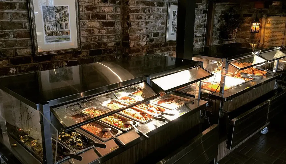 The image shows a buffet line with various dishes displayed under heat lamps in a room with brick walls and a framed picture