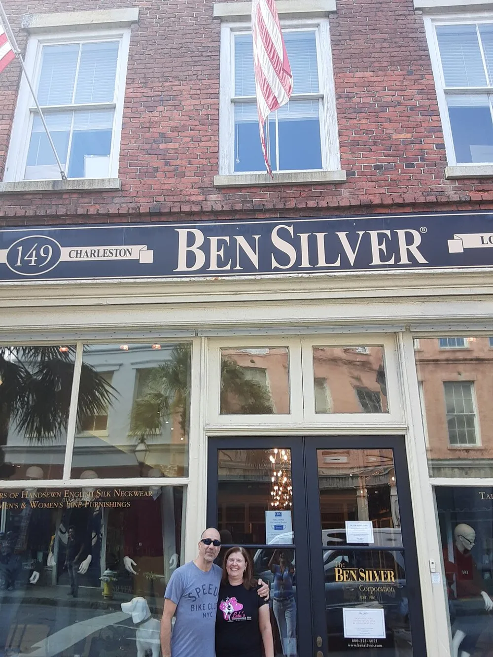 A man and a woman stand smiling in front of the Ben Silver storefront on a sunny day with American flags hanging above them