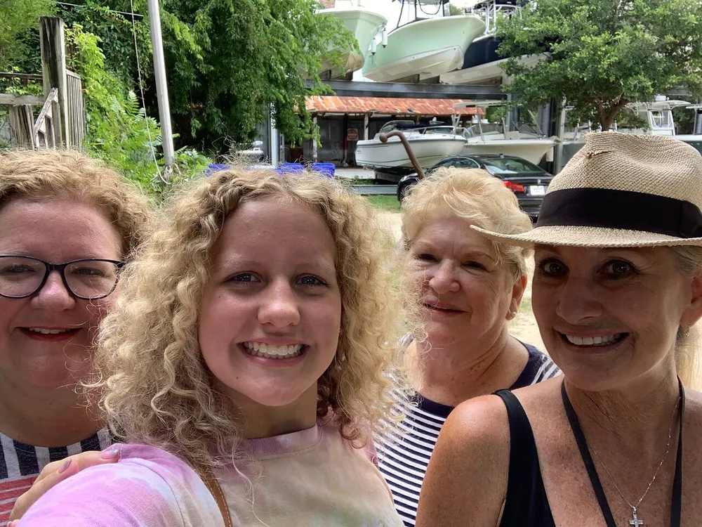Four smiling women are taking a close-up group selfie with greenery and stored boats in the background