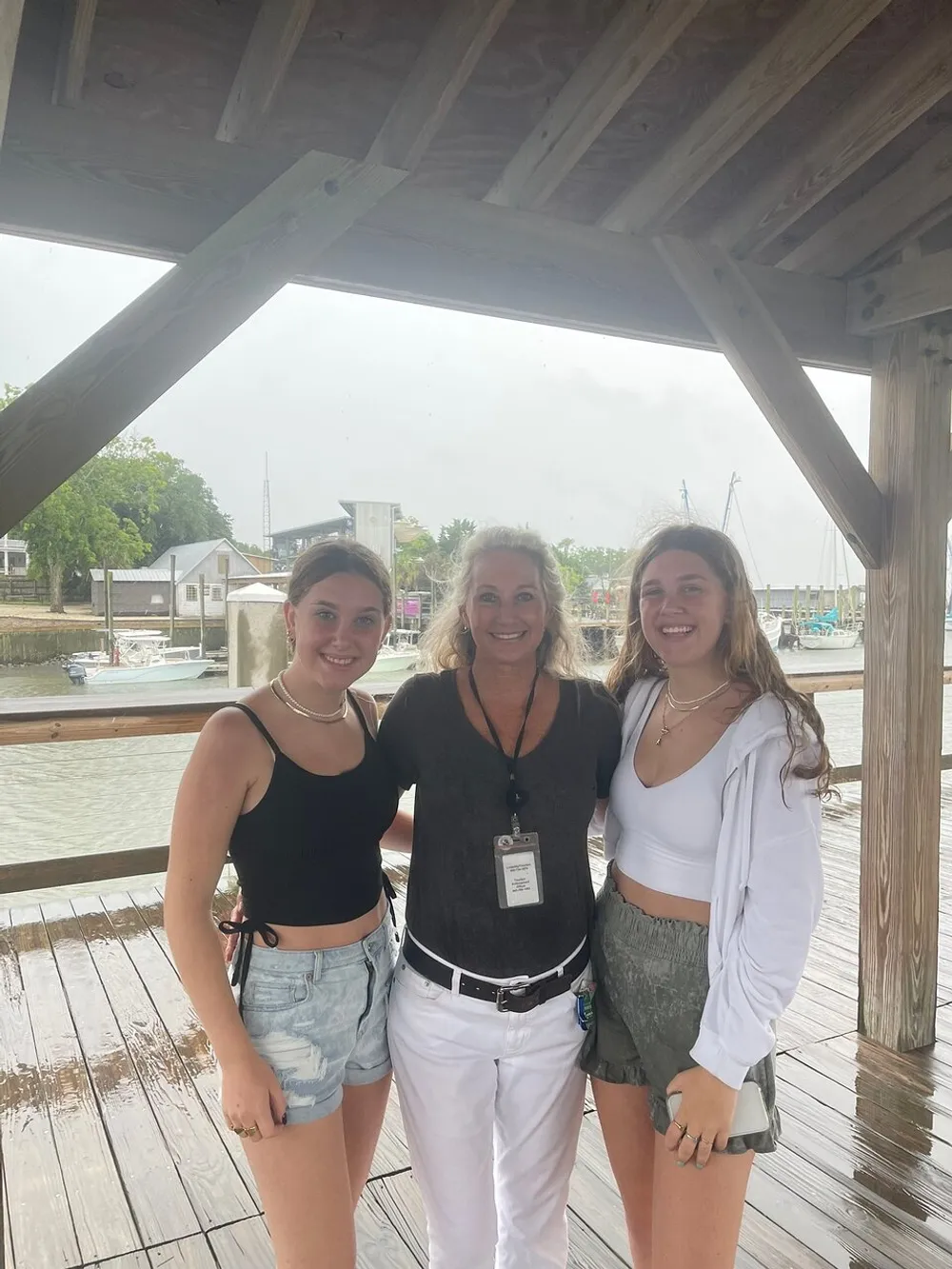 Three smiling people are standing under a wooden shelter by a marina on a cloudy day