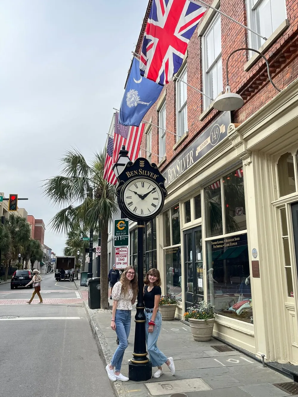 Two individuals are smiling and posing in front of a clock post on a sidewalk with flags hanging above them and a store named Ben Silver in the background