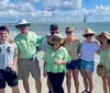 A group of seven people some wearing sun hats are smiling for a photo on a sunny beach with a lighthouse in the background