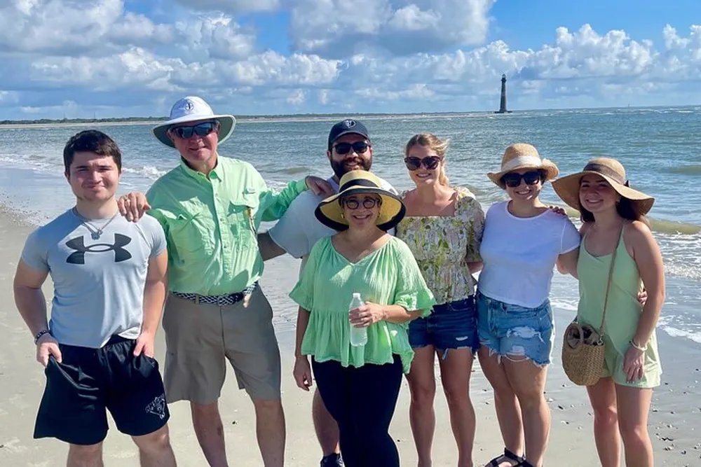 A group of seven people some wearing sun hats are smiling for a photo on a sunny beach with a lighthouse in the background