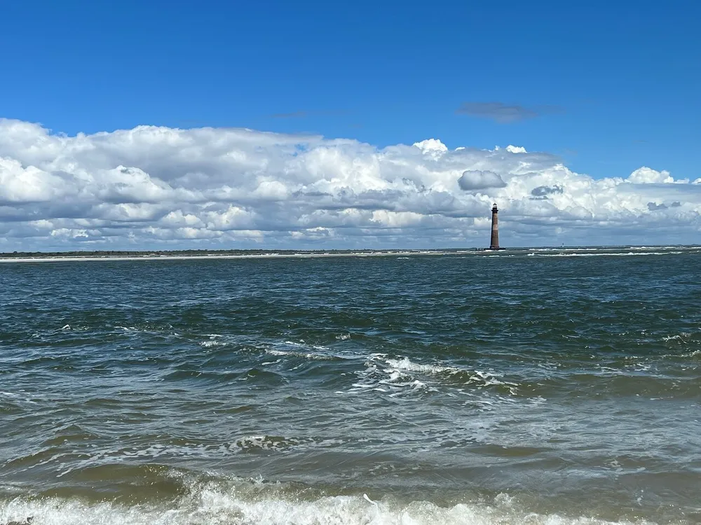 The image shows a tumultuous sea with choppy waves under a sky filled with billowing clouds anchored by a distant lighthouse standing tall near the horizon