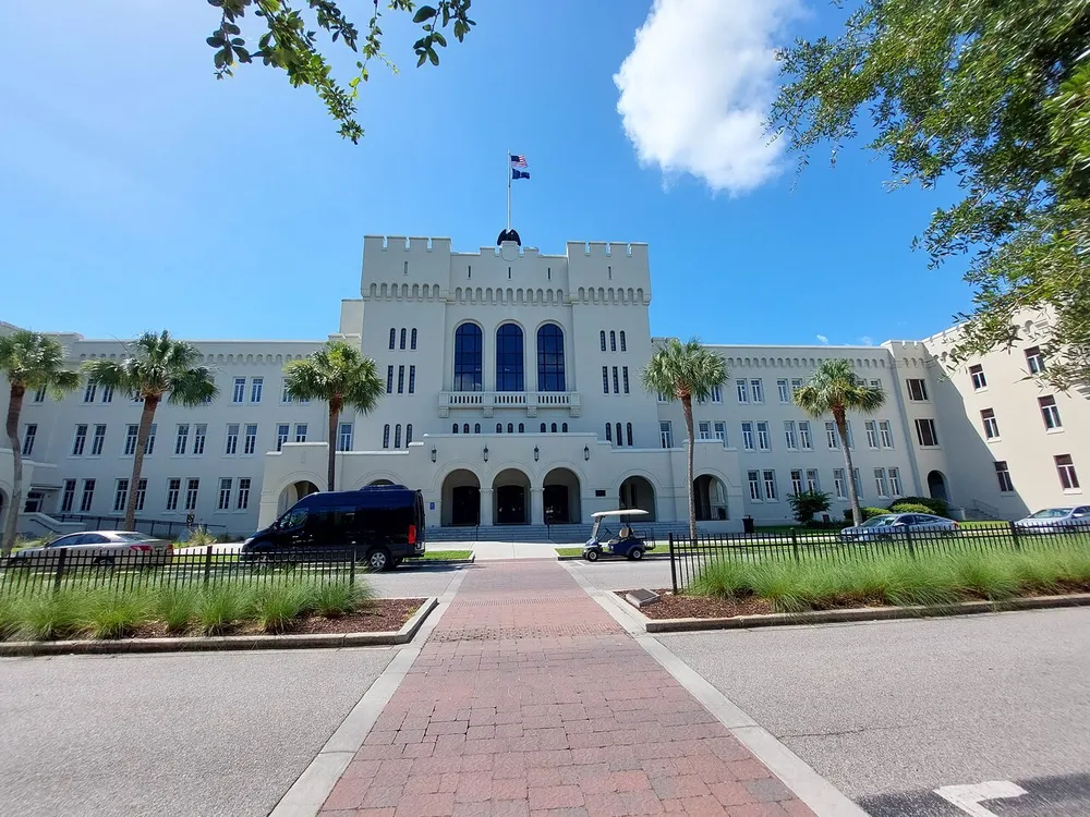 The image shows a large castle-like building with a central arched entrance flanked by rows of windows and palm trees under a bright blue sky with a few clouds