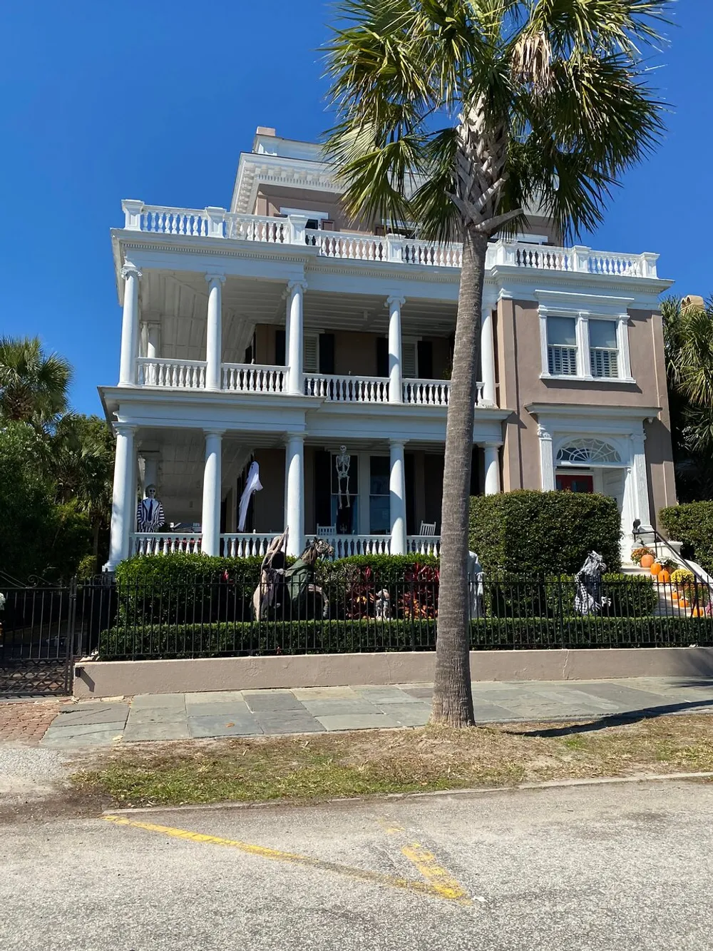 The image shows a grand multi-story house with double-decker porches adorned with white columns set behind a black iron fence with Halloween decorations visible all under a clear blue sky