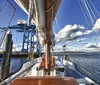 A two-masted schooner sails on calm blue waters under a clear sky passing beneath a large bridge in the background