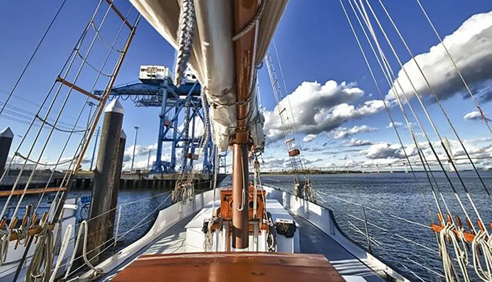 The image shows the deck of a tall ship with its rigging looking towards the bow with industrial cranes and a partly cloudy blue sky in the background