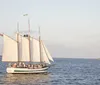A two-masted schooner sails on calm blue waters under a clear sky passing beneath a large bridge in the background
