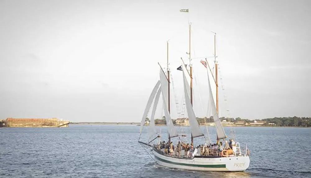 A classic schooner with its sails partially unfurled glides over the water with passengers on board against a backdrop of a distant shoreline