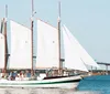 A two-masted schooner sails on calm blue waters under a clear sky passing beneath a large bridge in the background