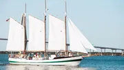 A two-masted schooner sails on calm blue waters under a clear sky, passing beneath a large bridge in the background.