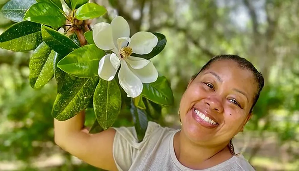 A joyful woman is holding a branch with a large white flower smiling brightly outdoors among the greenery