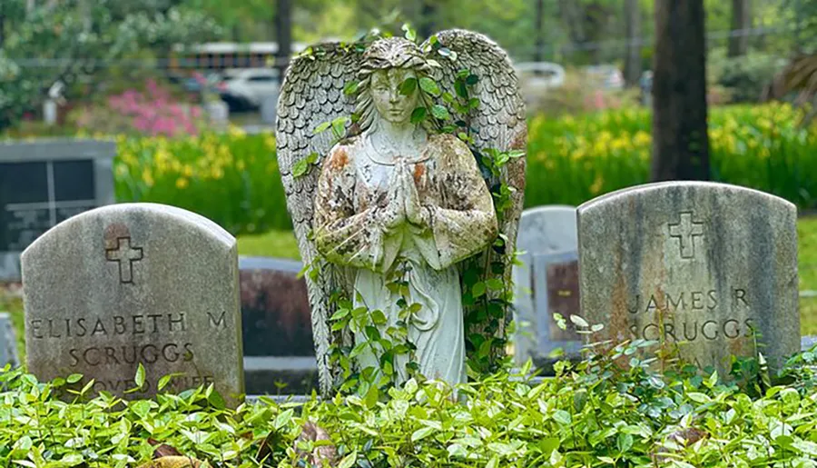 An angel statue with folded hands stands between two gravestones in a cemetery.