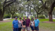 A group of six people is posing for a photo on a path lined with majestic oak trees draped with Spanish moss, with a large white house in the background.