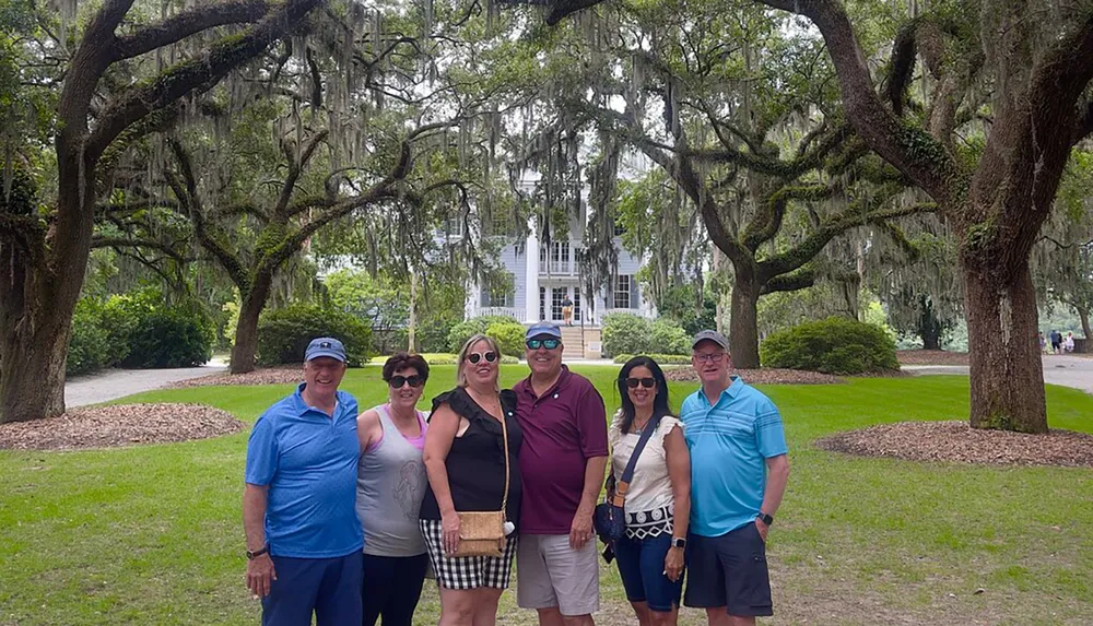 A group of six people is posing for a photo on a path lined with majestic oak trees draped with Spanish moss with a large white house in the background