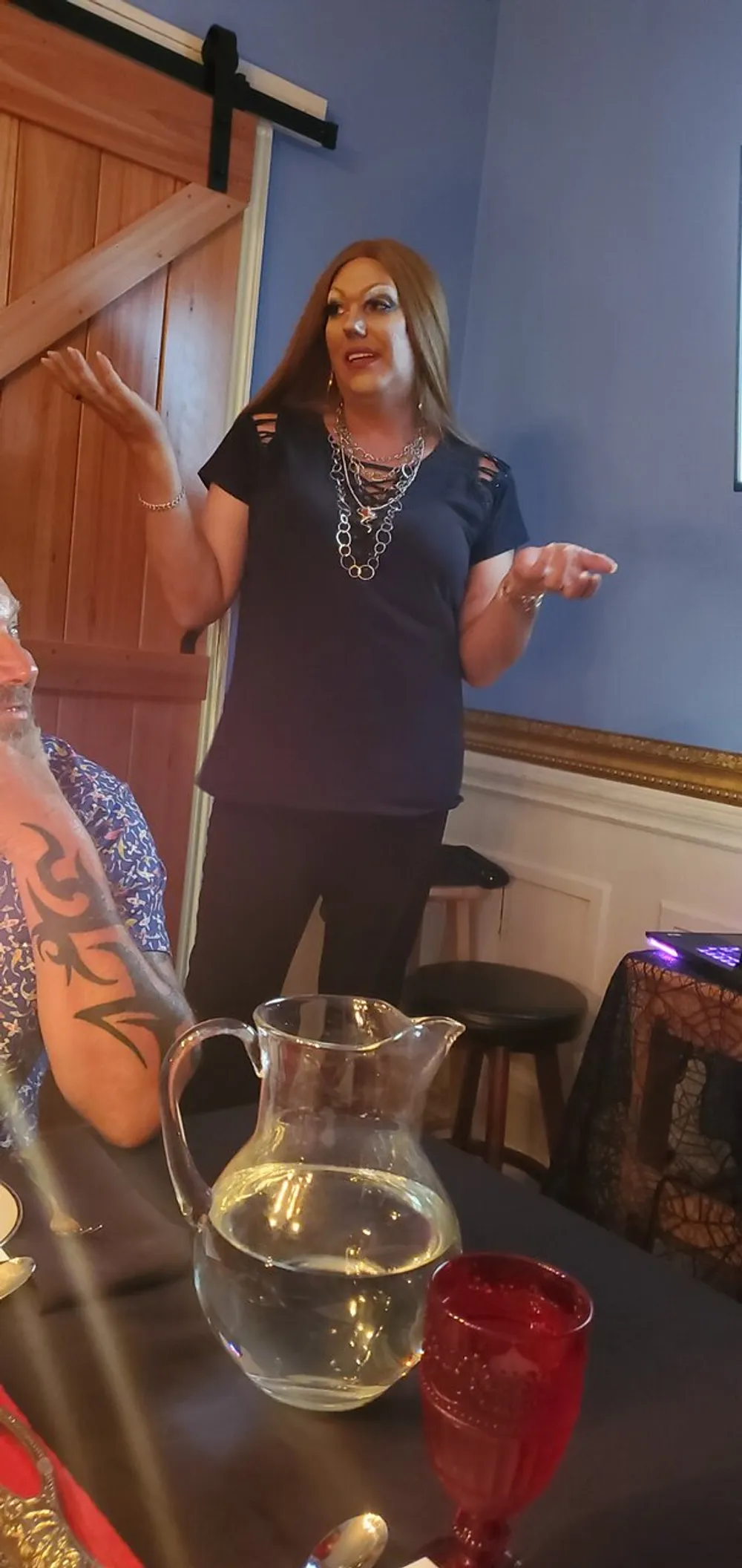 A person is standing and gesturing while speaking in a room with a water pitcher and red glass on a table and part of another person with a tattoo is visible in the foreground