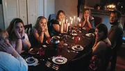 A group of surprised people sits at a dimly lit dinner table with lit candles, appearing to react to an unexpected event.