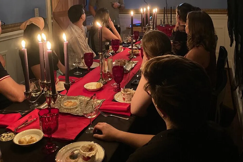Guests are gathered around a festively set dinner table with lit candles red accents and elegant tableware participating in a dimly lit intimate gathering