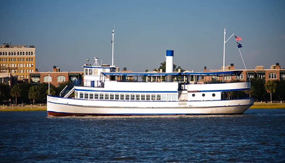 A vintage-style passenger ferry is cruising near the coastline against a backdrop of urban buildings under a clear sky