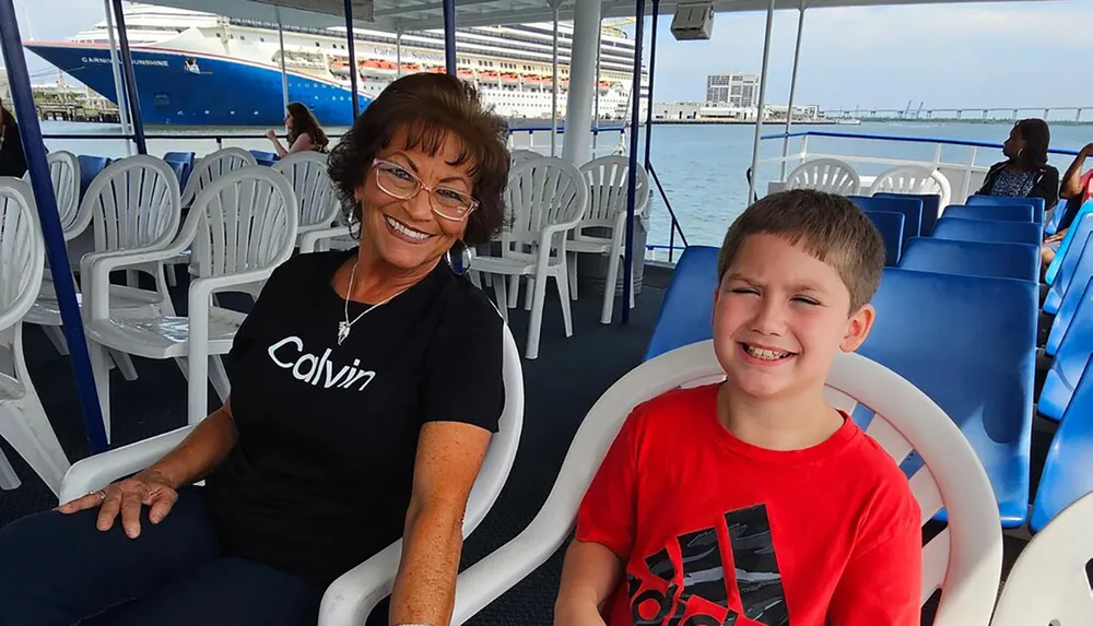 A smiling woman and a young boy are seated on a boat with a view of a large cruise ship in the background