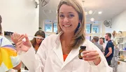 A woman in a white coat holds a dropper and a small bottle with a smile, as others engage in activities in the background of a brightly lit room.