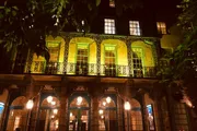 An illuminated building at night with intricate ironwork on the balcony and a warm yellow light casting a welcoming glow from its windows.