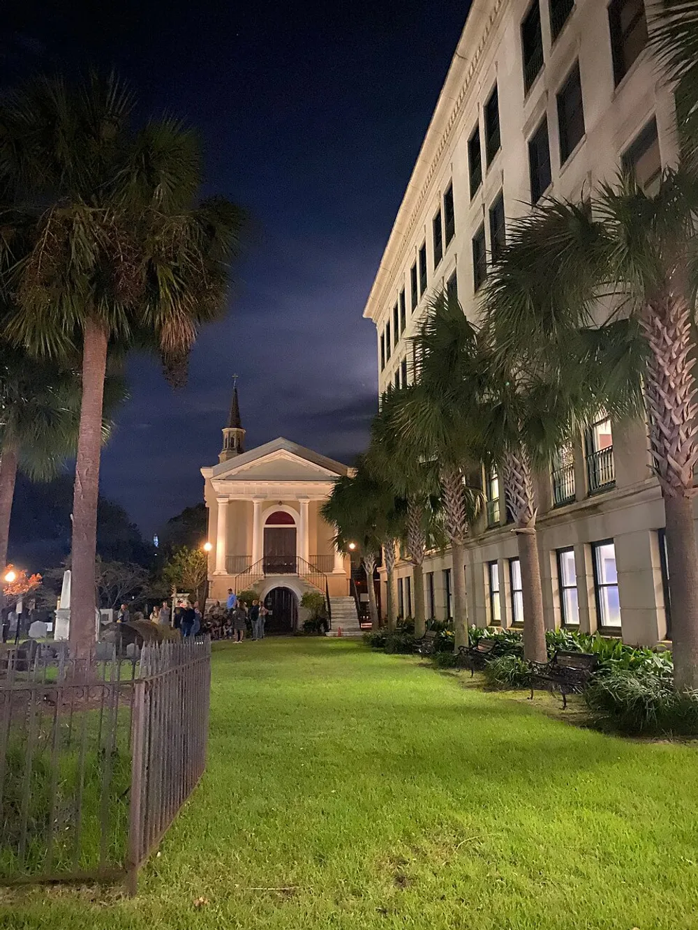 A nighttime view of a grand historic building possibly a church with people milling about flanked by tall palm trees and a deep blue sky