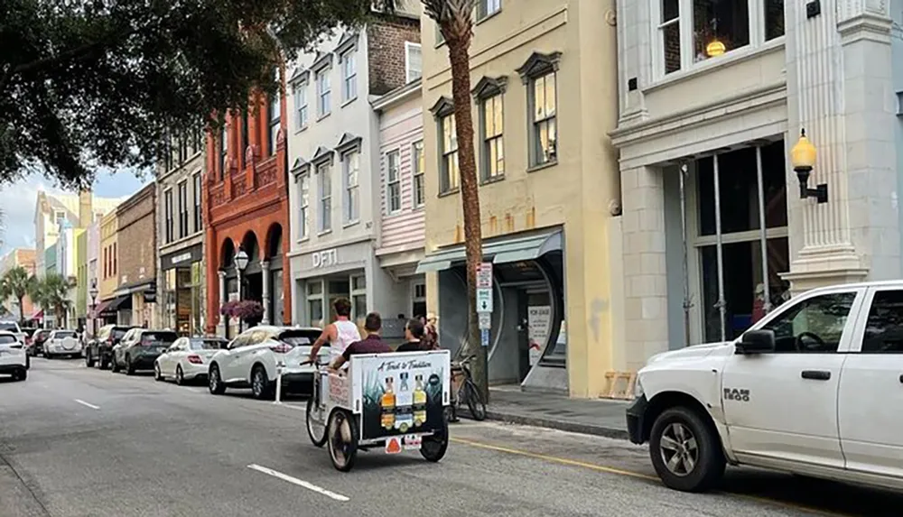 A pedicab advertising spirits is being ridden by a driver with two passengers on a street lined with colorful buildings and parked cars