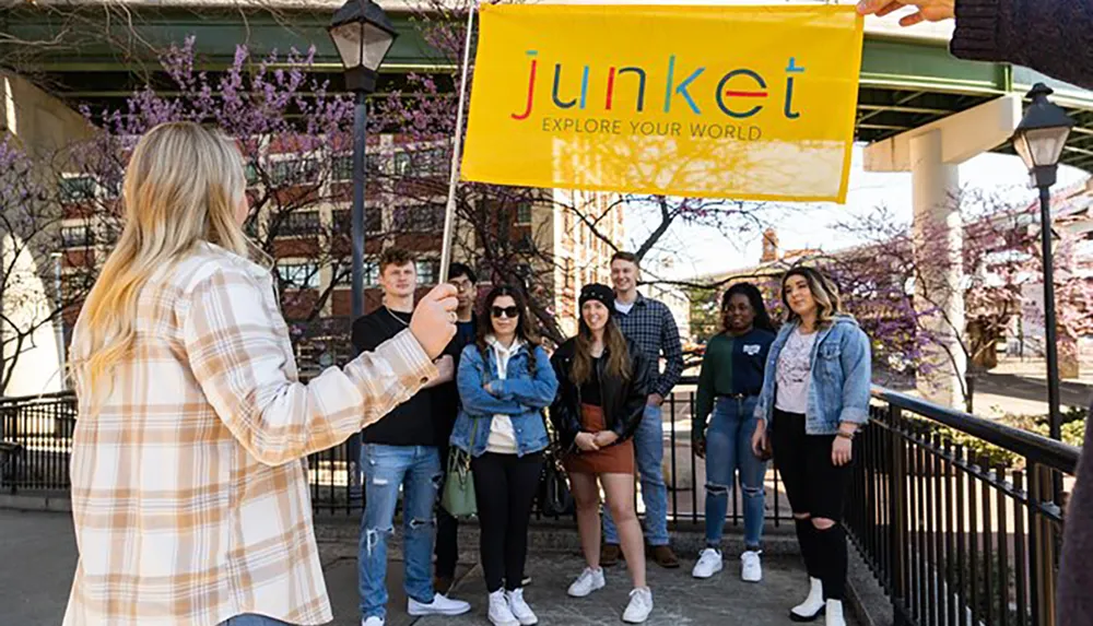 A group of smiling people pose for a photo under a yellow banner with the word junket while a woman in the foreground takes the picture with her smartphone