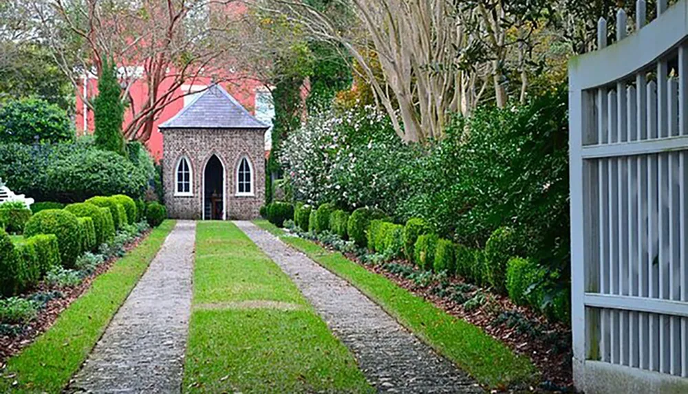 A quaint stone building with a pointed arched doorway sits at the end of a landscaped path flanked by neatly trimmed hedges and a wooden gate to the right