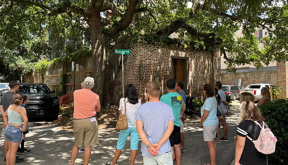 A group of people is gathered on a shady street corner near a brick structure with a street sign reading Bedons Alley