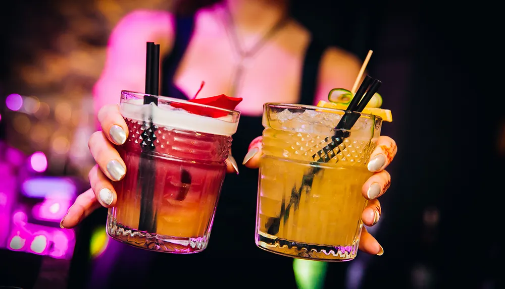 A person is holding two colorful cocktails against a blurred background of lights