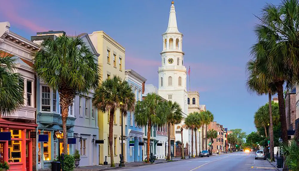 A tranquil city street at dusk lined with colorful buildings and palm trees leading the eye toward a tall white historical church tower against a soft twilight sky