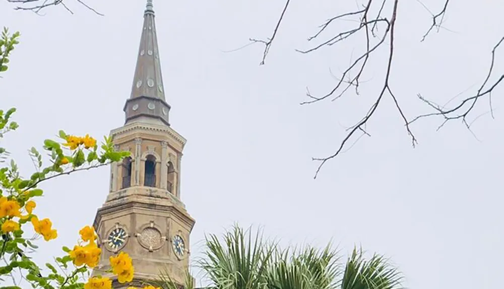 The image shows the top of a historic-looking church steeple with a clock framed by greenery and flowers in the foreground under a cloudy sky