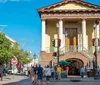 This image depicts a bustling street scene with pedestrians and a car in front of an elegant building with a sign reading Daughters of Confederacy under a clear blue sky