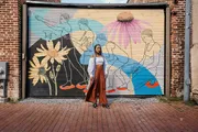 A woman stands in front of a colorful garage door mural featuring abstract human figures and sunflowers.