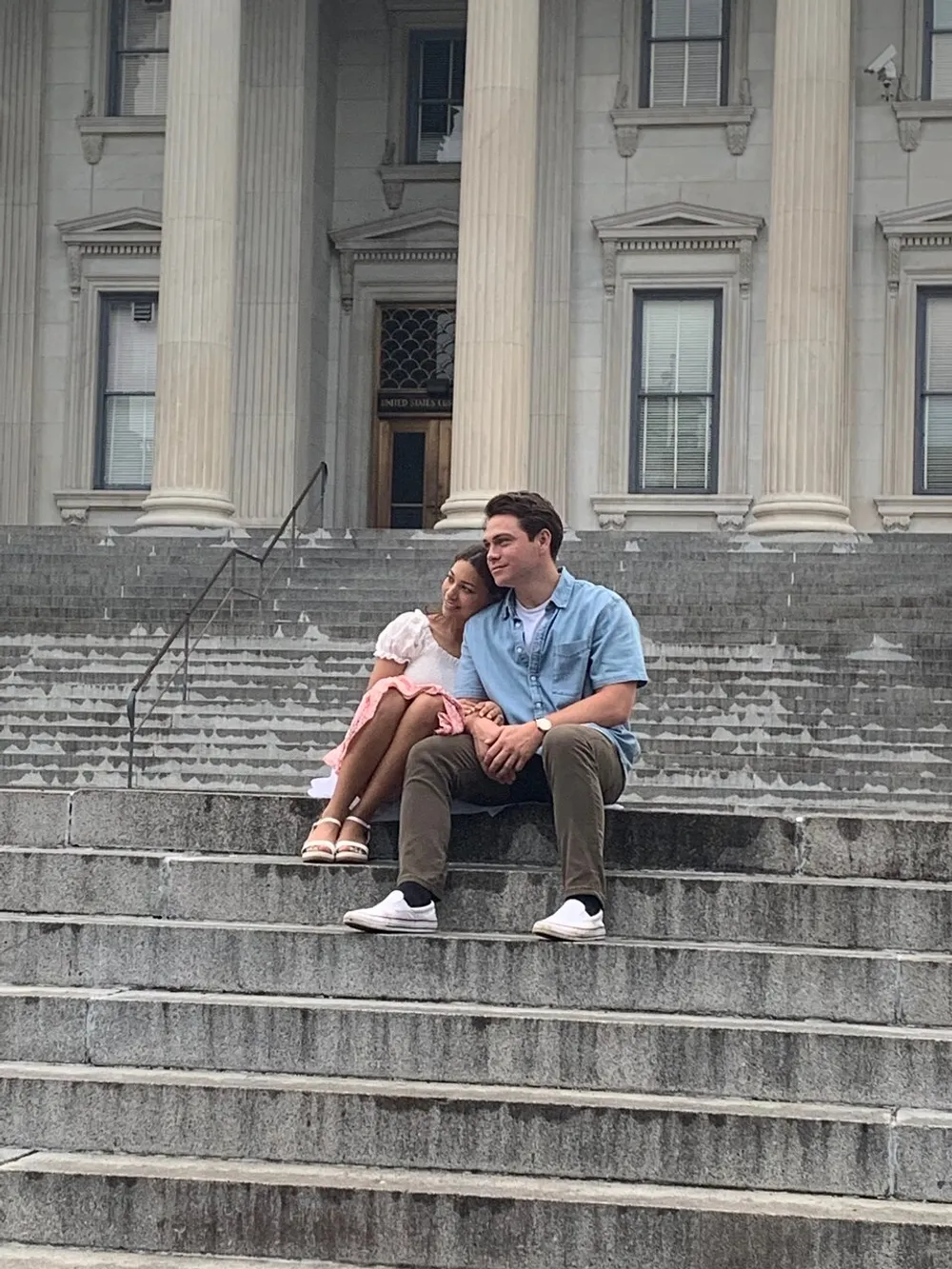 A couple is sitting closely together on the steps of a neoclassical building with one partner affectionately resting their head on the others shoulder