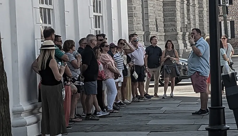 A group of people is attentively listening to a tour guide who is explaining something outdoors next to a building with white walls