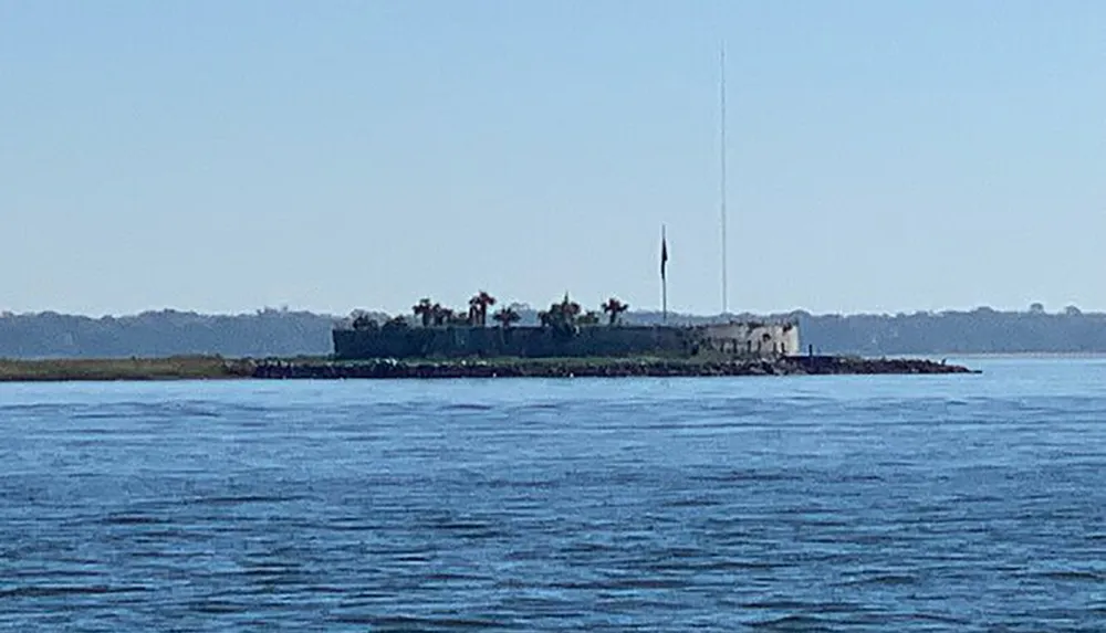 The image shows a small island with structures and palm trees viewed from across a body of water under a clear sky