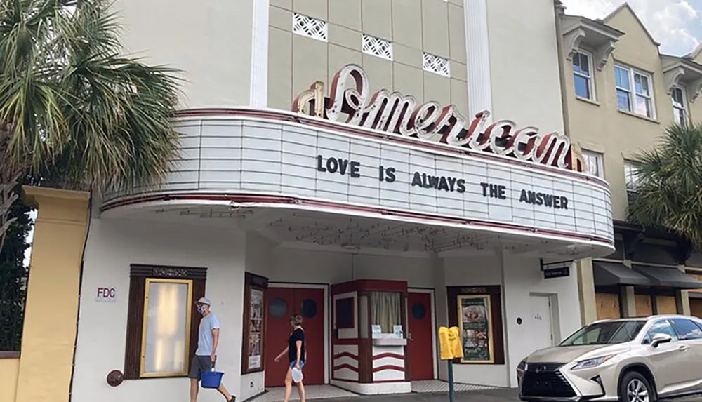 The image shows the front of a classic American theater with a marquee that reads LOVE IS ALWAYS THE ANSWER as people walk by and a car is parked in front