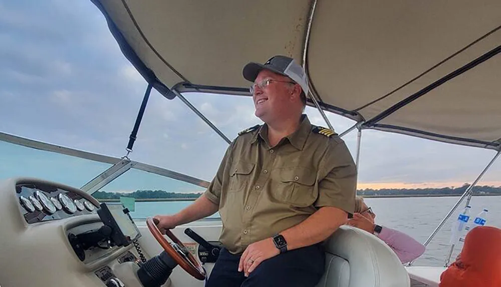 A person in a captains uniform is smiling and steering a boat with water and twilight sky in the background