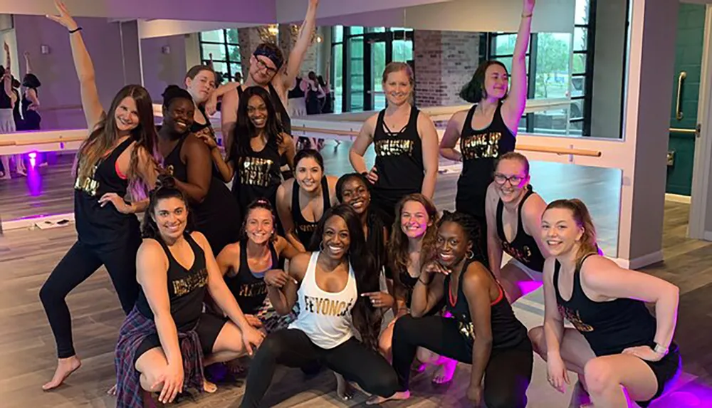 A group of smiling people in dance or workout attire are posing together in a studio with mirrors and a ballet barre in the background
